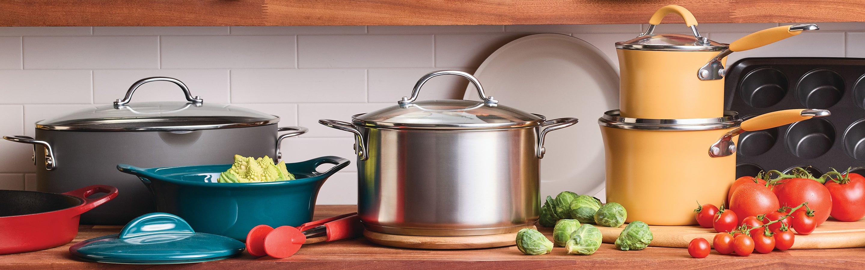 Various pots, pans, and kitchenware are arranged on a wooden countertop with fresh vegetables, tomatoes, and Brussels sprouts in a kitchen setting.