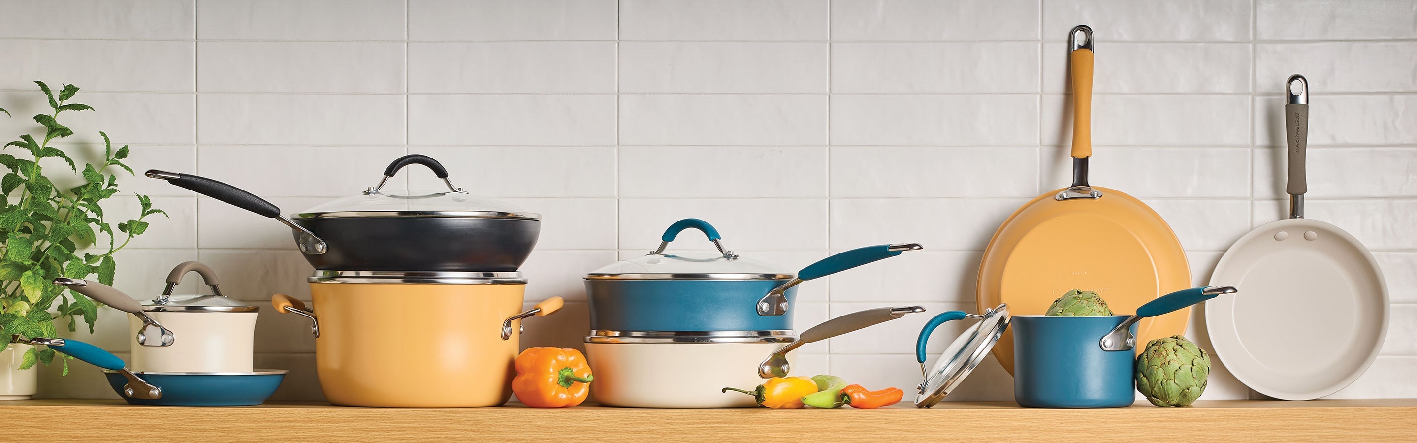 A set of various saucepans, pots, and frying pans with blue, yellow, and white exteriors arranged on a wooden countertop in front of a white tiled wall.