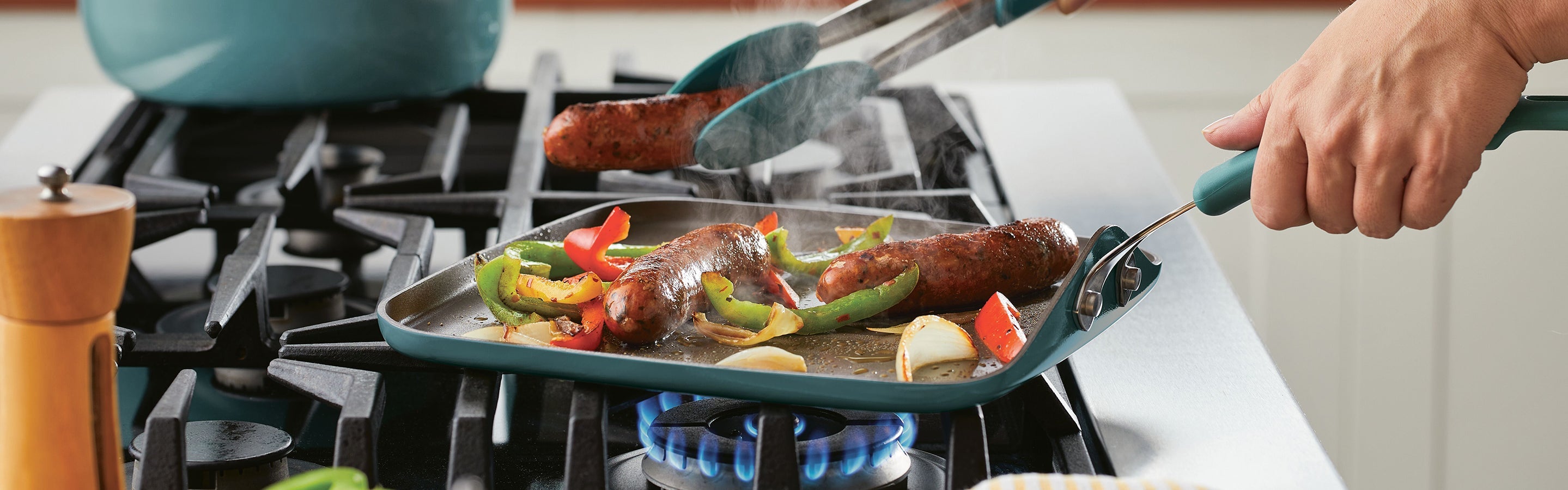 A person uses tongs to cook sausages and sliced bell peppers on a stovetop grill pan over a blue gas flame.