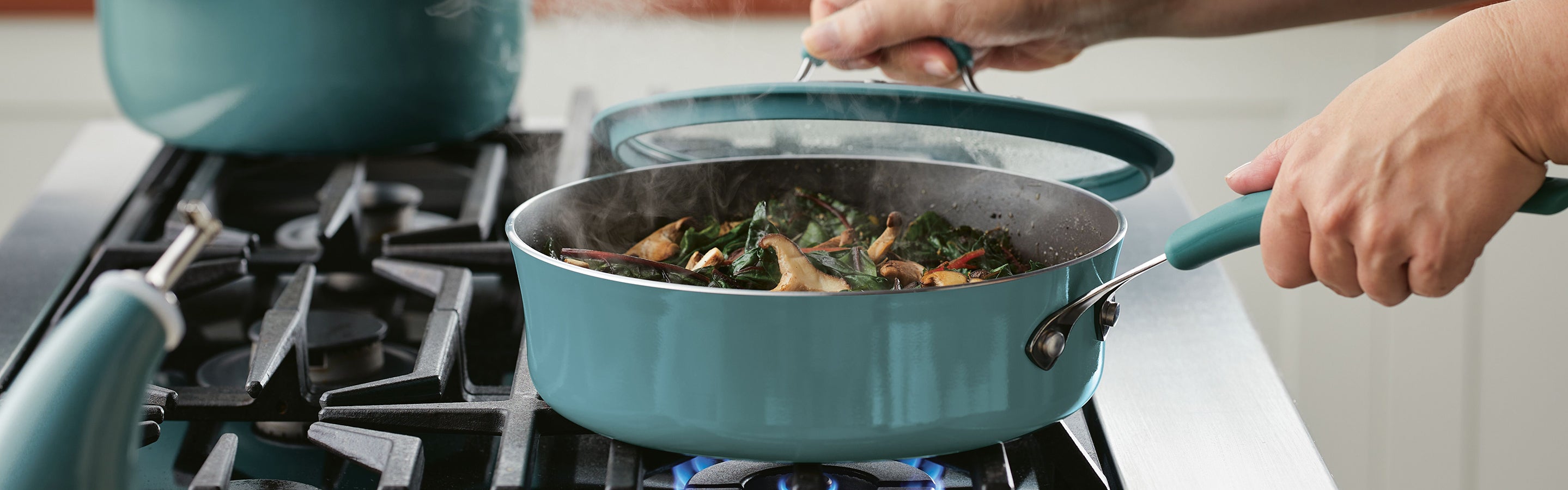 A person lifts the lid from a teal saucepan on a gas stove, revealing steaming greens and mushrooms cooking inside.