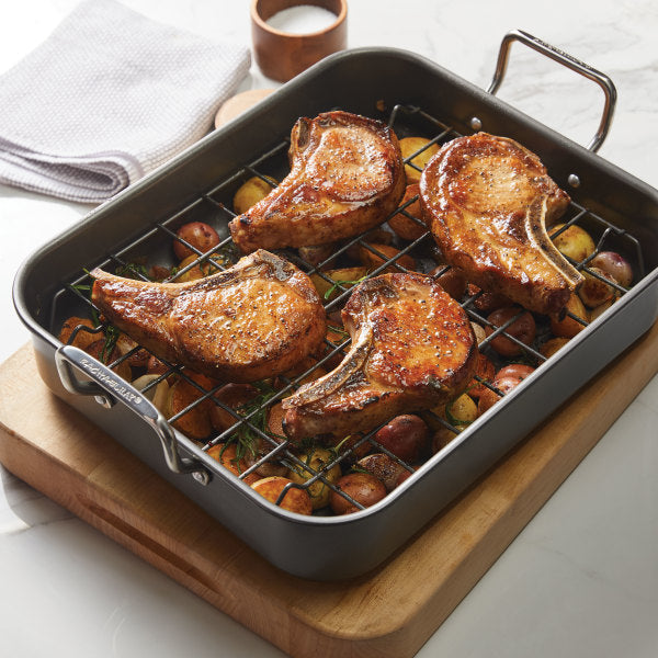 Pork chops roasting on the reversible rack of a 16.5-Inch x 13.5-Inch Nonstick Steel Roaster, placed over seasoned vegetables.