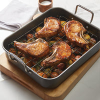 Pork chops roasting on the reversible rack of a 16.5-Inch x 13.5-Inch Nonstick Steel Roaster, placed over seasoned vegetables.