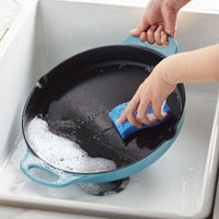 Hands scrubbing a black 14-Inch NITRO Cast Iron Skillet with a blue sponge in a sink, ensuring it's induction ready and oven safe for your culinary adventures.