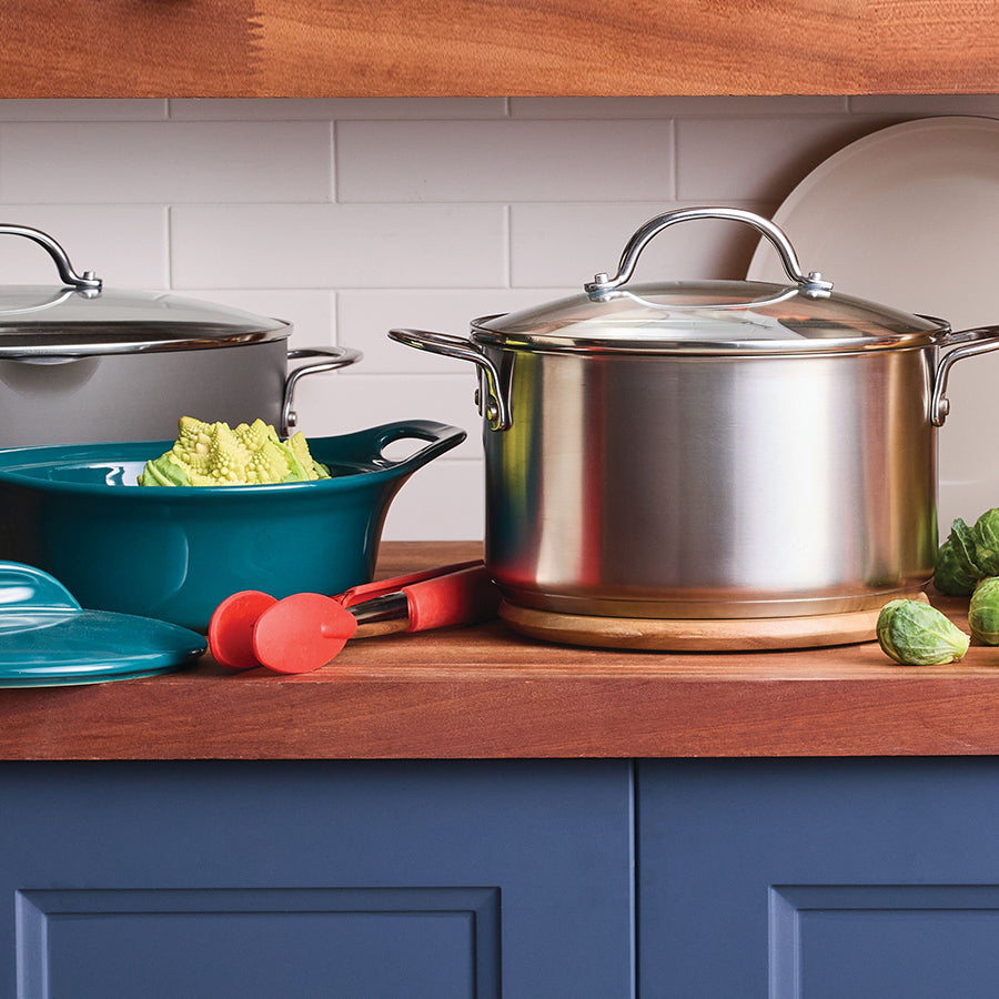 A curated mix of Rachael Ray collections on a wooden countertop. It showcases a polished stainless steel stockpot with a glass lid alongside an agave blue ceramic pot filled with romanesco broccoli, paired with red kitchen tongs and fresh Brussels sprouts against a white subway tile backsplash.