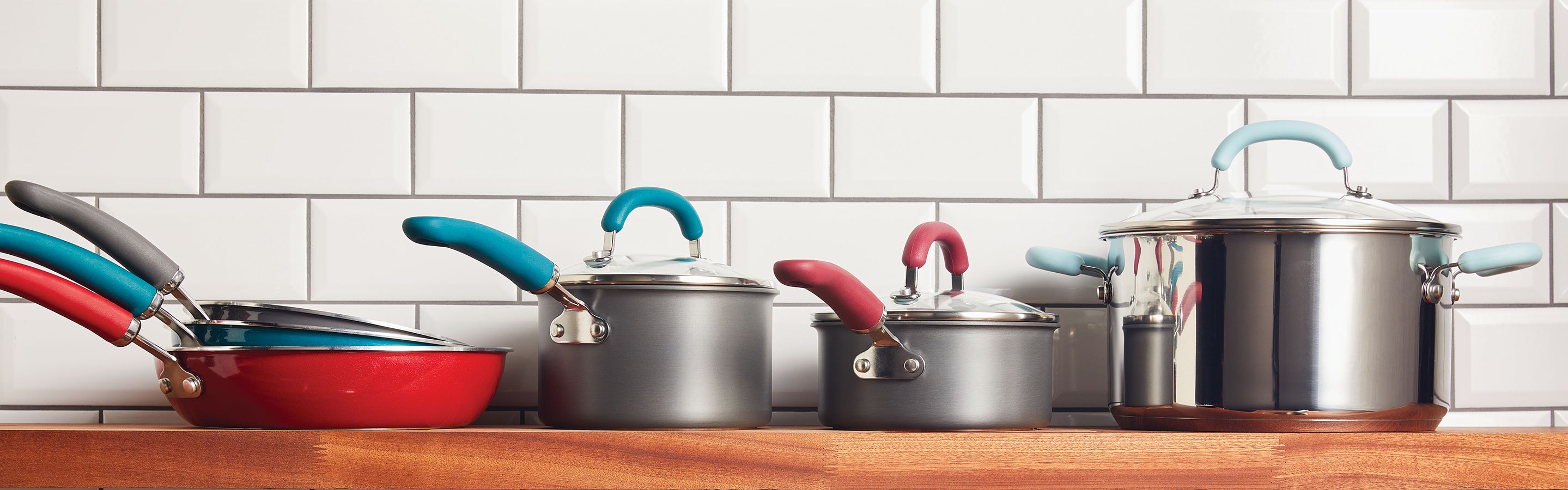 A variety of Rachael Ray Create Delicious cookware is displayed on a wooden shelfagainst a white subway tile backsplash. The collection includes pots and pans in various sizes with shimmering silver exteriors and colorful handles.