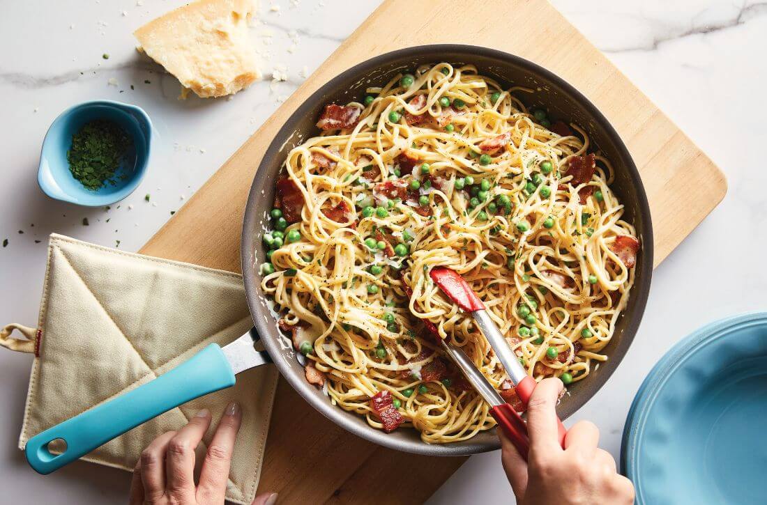 Egg Tagliatelle Carbonara with peas and spring onions being tossed with red tongs in a blue Rachael Ray Cook + Create nonstick saute pan.
