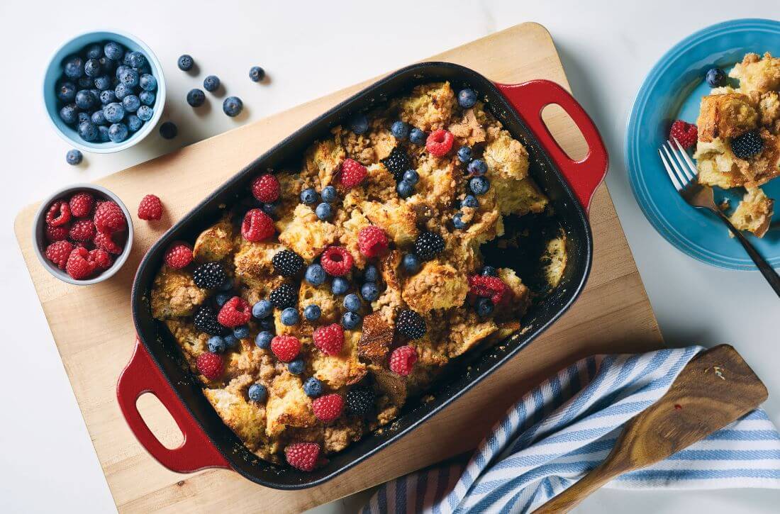 A French toast casserole topped with fresh blueberries, raspberries, and blackberries, served in a red Rachael Ray Nitro Cast Iron roasting pan on a wooden cutting board.