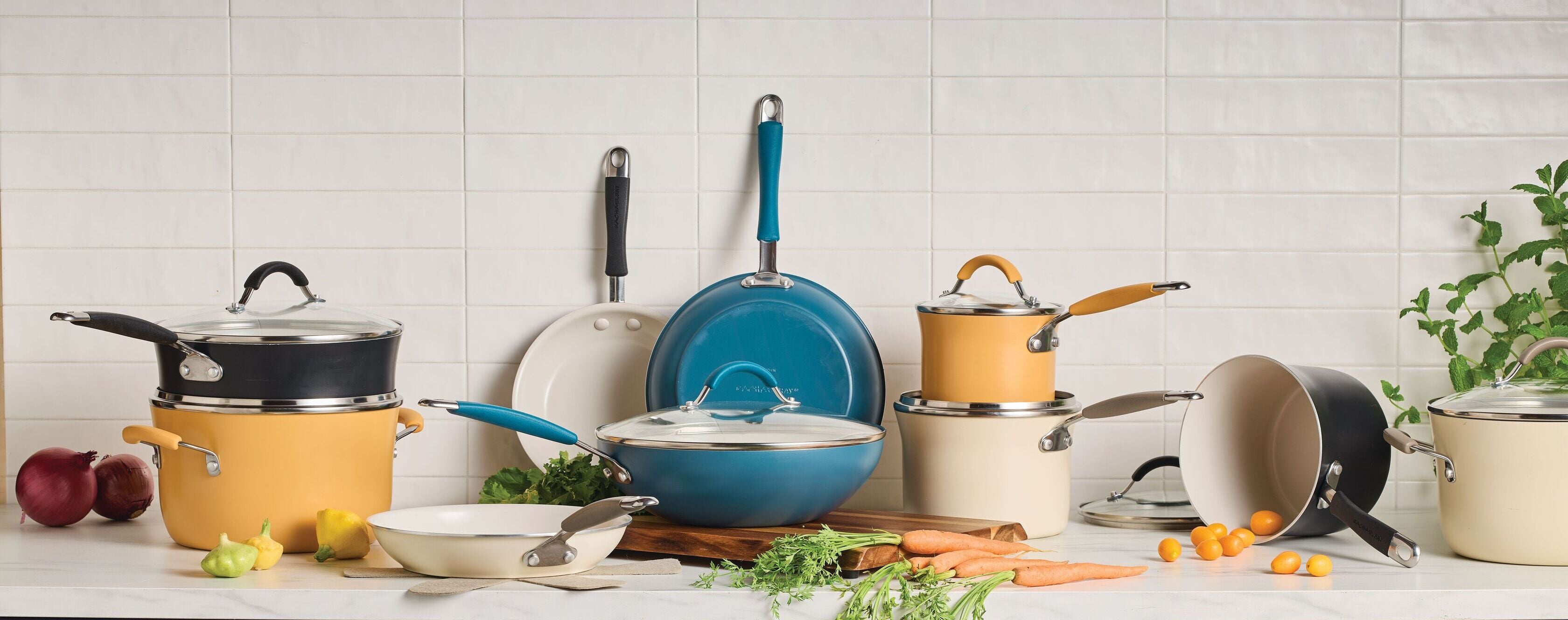 An assortment of Rachael Ray Cucina Ceramic cookware in blue, yellow, cream, and black colors arranged on a marble countertop with a white tile backsplash.