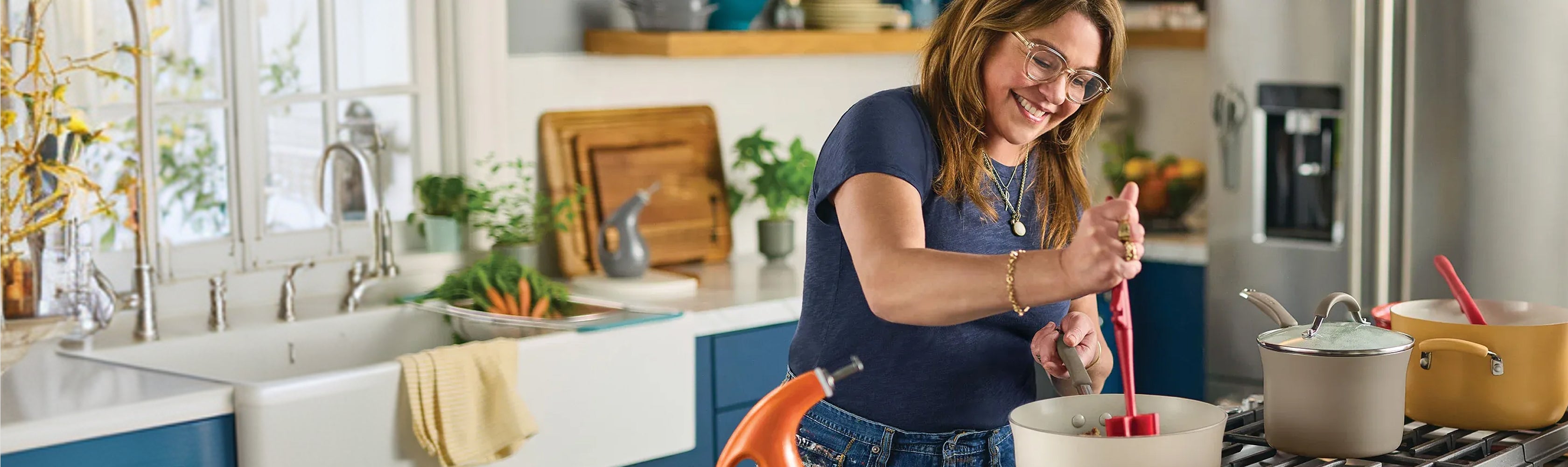 Rachael Ray smiling while stirring a pot in a modern kitchen, with a variety of colorful Cucina ceramic cookware and fresh ingredients on the counter.