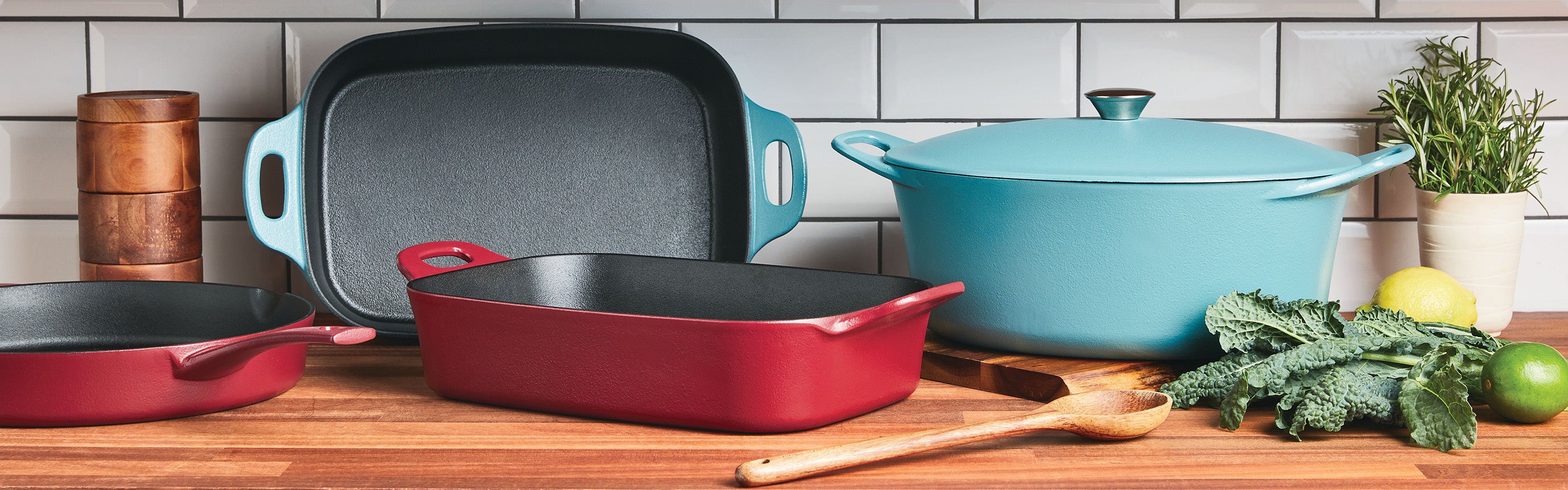 Rachael Ray NITRO Cast Iron collection display is arranged on a wooden countertop against a white subway tile backsplash. An agave blue Dutch oven and griddle sit alongside a red rectangular baker and skillet, accompanied by a wooden spoon, fresh herbs, and citrus.