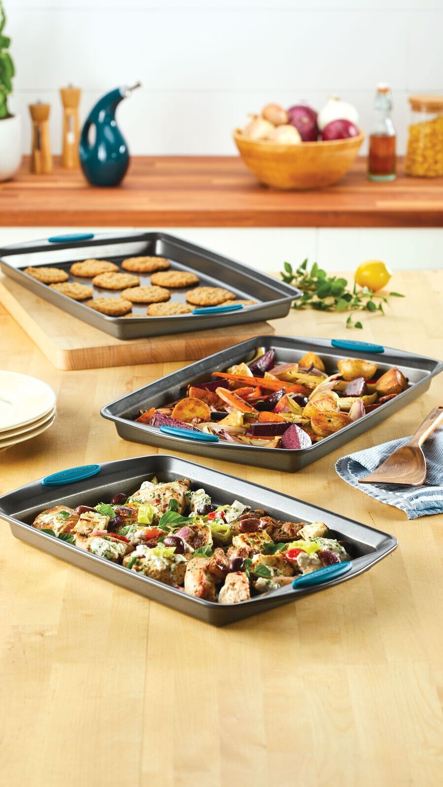 A three-piece set of Rachael Ray sheet pans with marine blue grips, featuring roasted vegetables, chicken, and freshly baked cookies on a wooden tabletop.