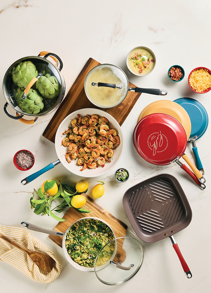 A variety of Rachael Ray cookware is arranged on a wooden countertop and cutting board, featuring a white saute pan with shrimp, a square grill pan, and a red circular pan. The collection is complemented by fresh ingredients like artichokes, lemons, and bowls of shredded cheese and toppings against a light marble background.