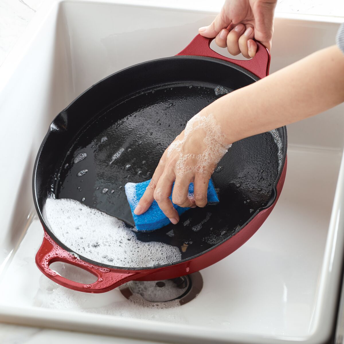 Hand washing a 14-Inch NITRO Cast Iron Skillet with a blue sponge in a white sink.