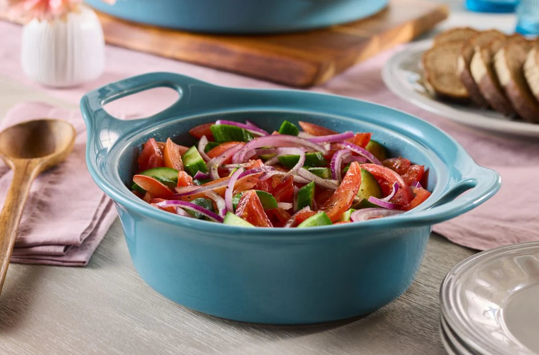 A Tomato, Cucumber, and Onion Salad served in an Agave Blue Rachael Ray Ceramic Au Gratin dish, set on a wooden table with sliced bread in the background.