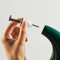 A hand carefully holds a disassembled stainless steel pour spout next to an EVOO Bottle 24-Ounce in green, ready for precision pouring.