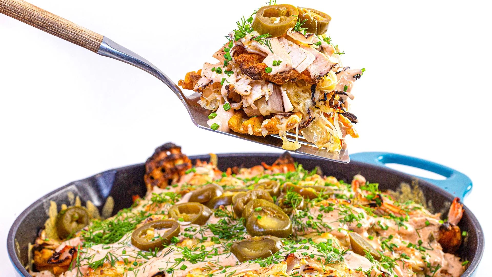 A metal spatula lifting a serving of Rachel's Reuben Nachos topped with sauerkraut, melted cheese, dressing, and sliced jalapeños from a blue Rachael Ray cast iron skillet.
