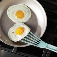 Two sunny-side-up eggs in a frying pan, with one being lifted by a spatula from the 10-Inch & 12-Inch Nylon Spatula Set.
