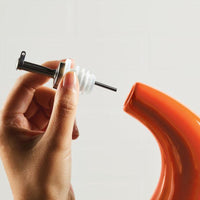 A hand holding a stainless steel pour spout beside an orange curved object, designed for precision pouring from the 24-Ounce EVOO Bottle.