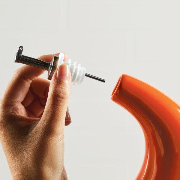 A hand holding a stainless steel pour spout beside an orange curved object, designed for precision pouring from the 24-Ounce EVOO Bottle.
