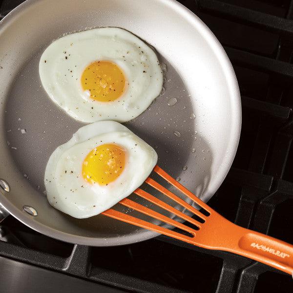 Two fried eggs in a pan with an orange spatula from the 10-Inch & 12-Inch Nylon Spatula Set.